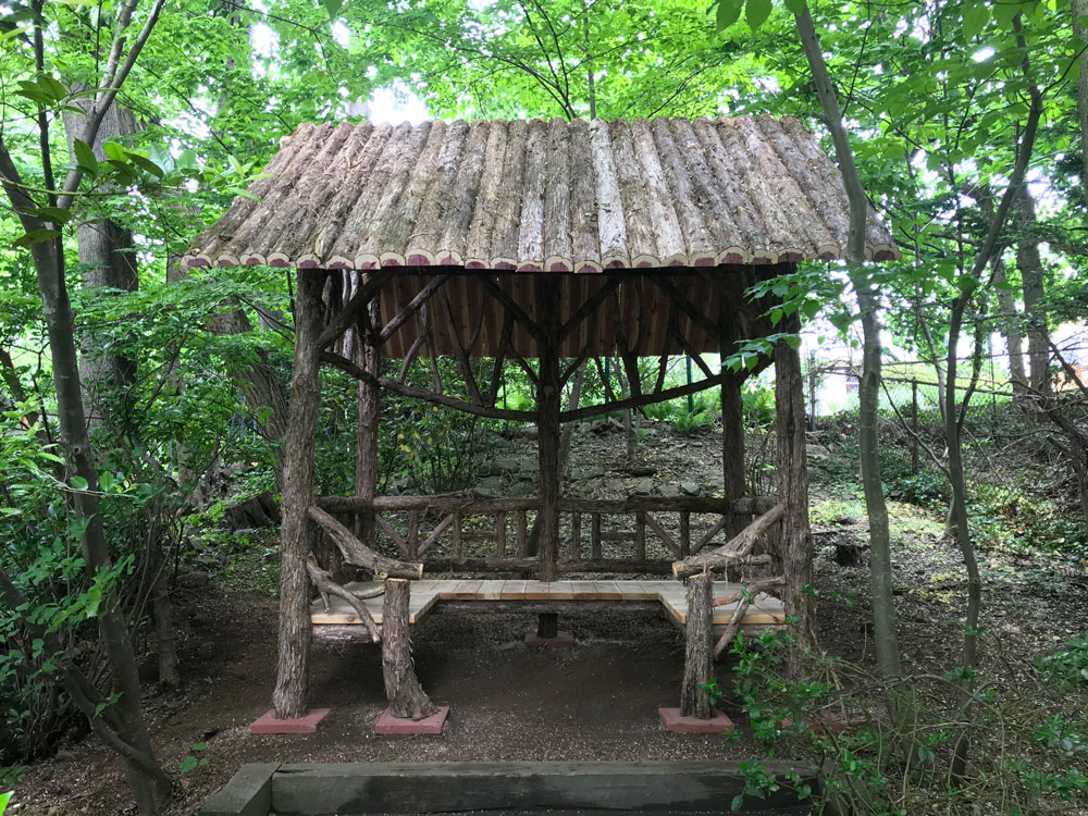 Rustic sitting shelter custom built using cedar trees and branches titled the Mayhew Shelter
