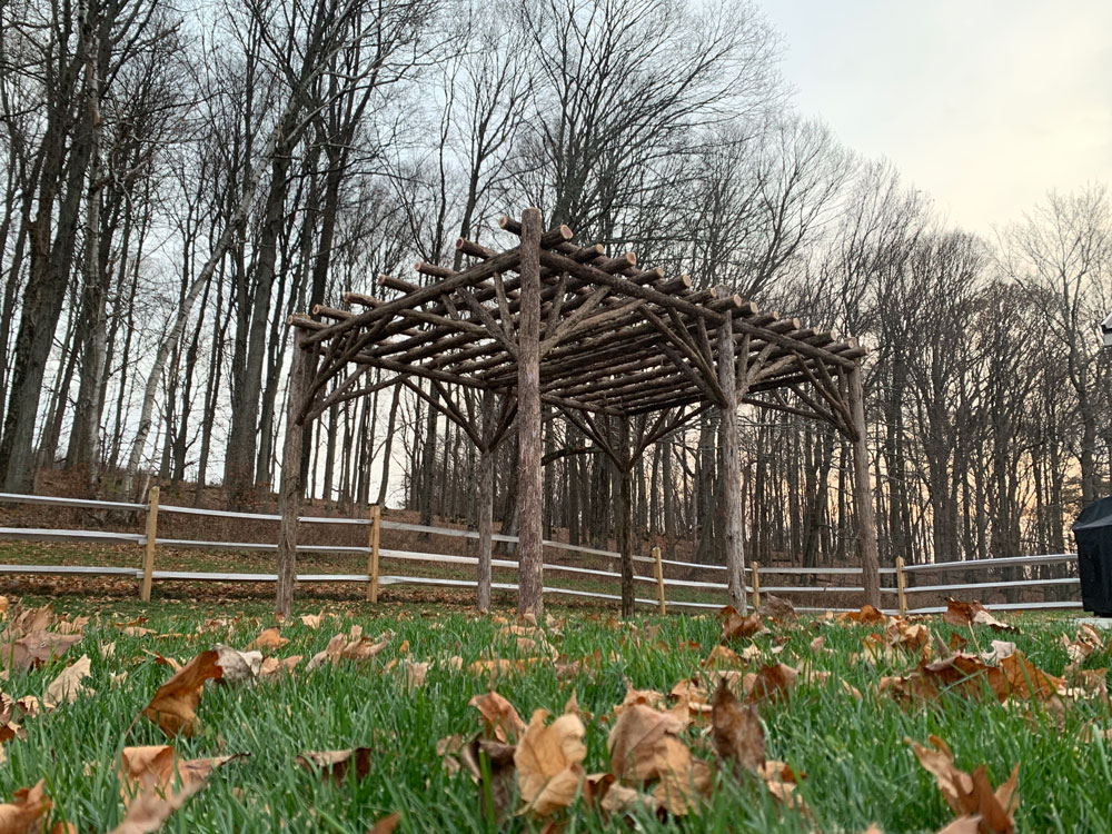 Rustic natural pergola made from bark-on cedar logs, twigs, and branches titled the Pine Plains Pergola