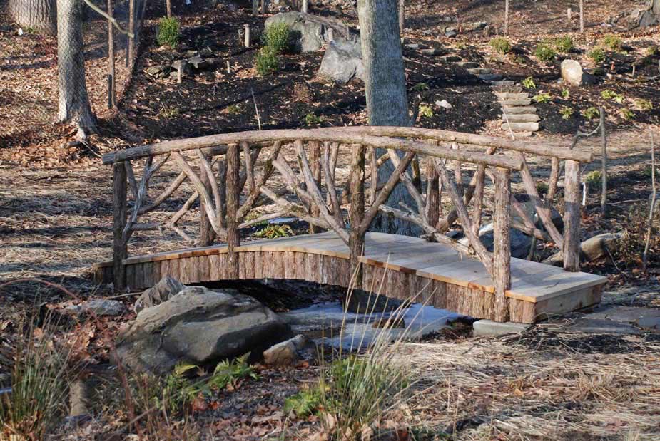Rustic bridge built over a steam using bark-on trees and branches titled the Grape Bridge